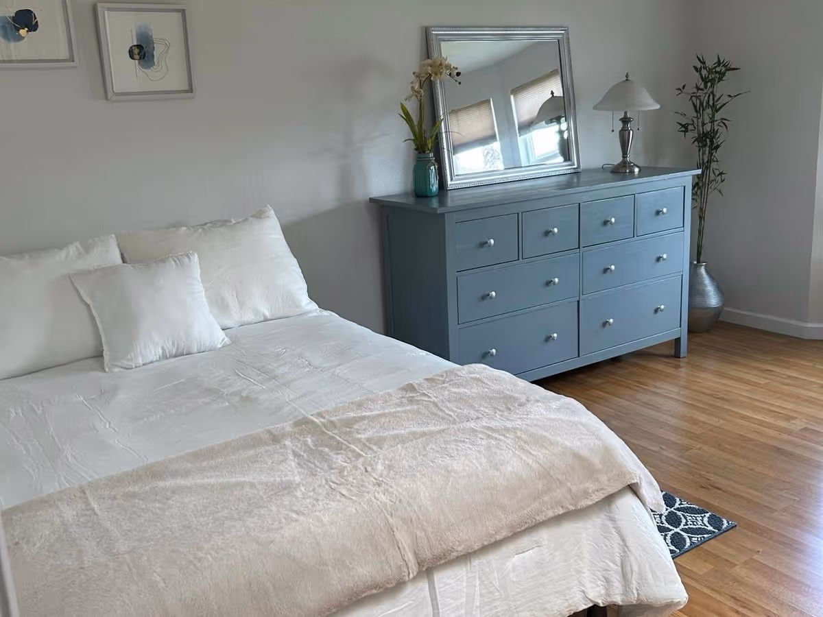 A bedroom with a neatly made bed featuring white pillows and a beige blanket. Next to the bed is a blue dresser with multiple drawers, topped with a mirror, a lamp, and a vase with flowers. The room has wooden flooring and a potted plant in the corner.