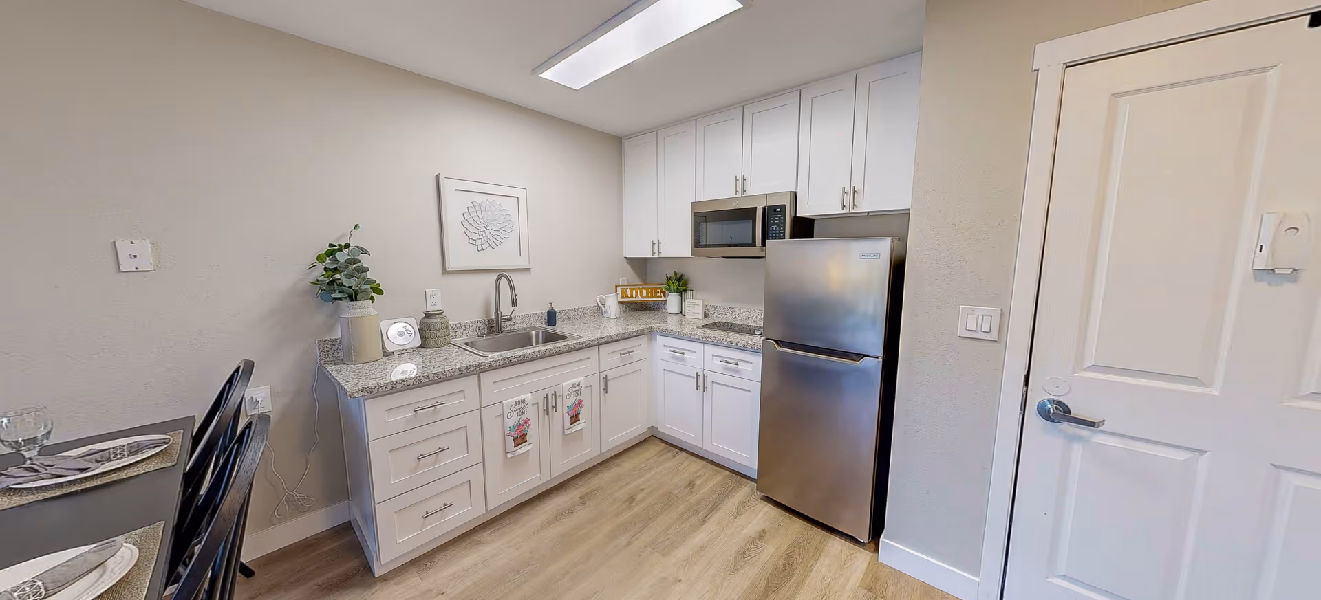 A modern kitchen area with white cabinets, a granite countertop, a stainless steel refrigerator, and a built-in microwave. There is a sink with a faucet, decorative plants, and kitchen towels hanging on the cabinet doors. To the left, part of a dining table with place settings is visible. The floor is light wood, and the walls are painted light gray.