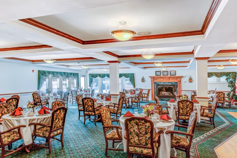 A spacious dining room with round tables covered in white tablecloths, each set with red napkins, glassware, and floral centerpieces. The chairs have floral upholstery. The room features green carpet with a patterned border, white walls with wooden trim, large windows with green valances, and a decorative fireplace with framed pictures above it.