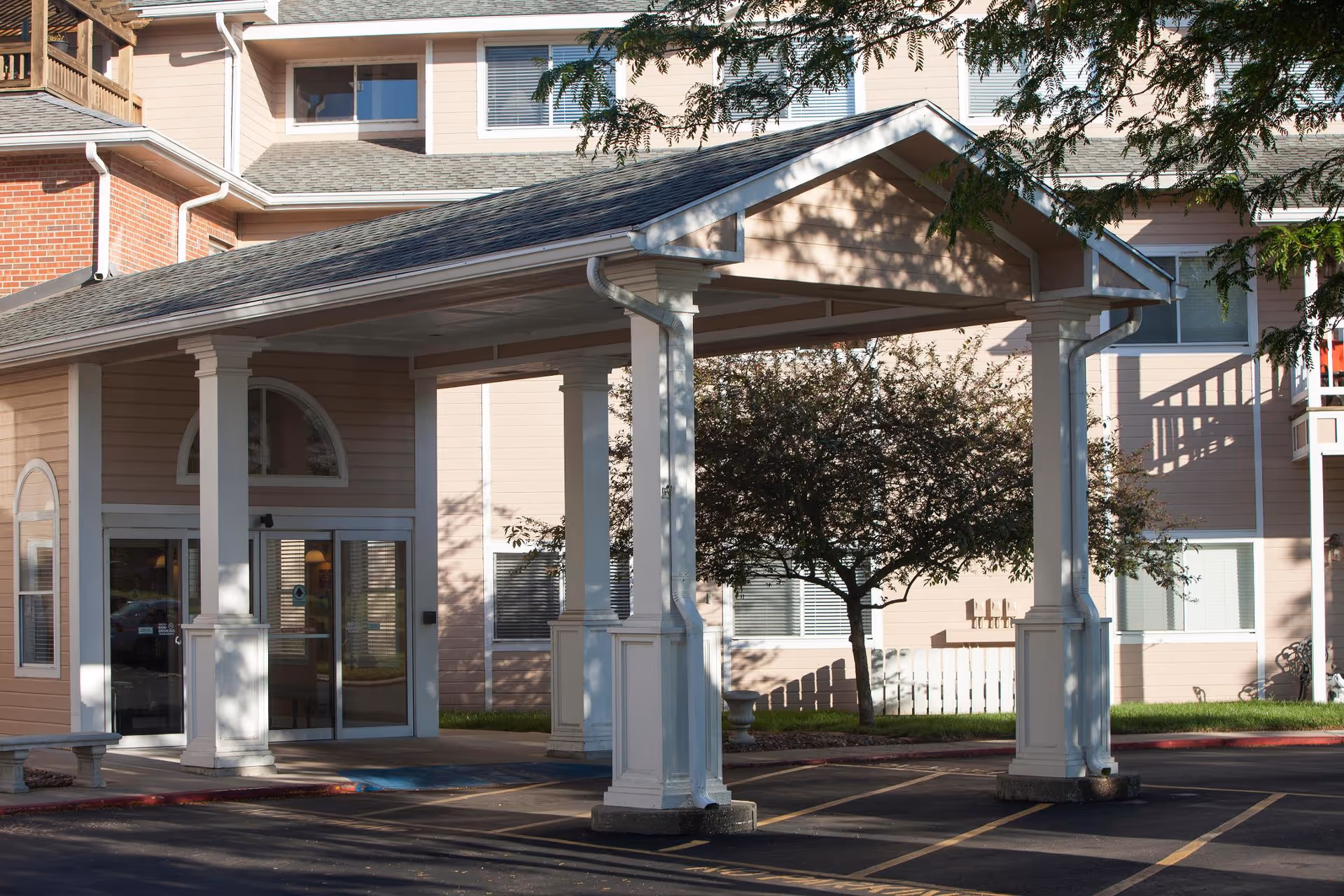 Entrance of a senior living facility with a covered drop-off area supported by white columns. The building has beige siding with white trim and several windows. There is a tree and some landscaping near the entrance.