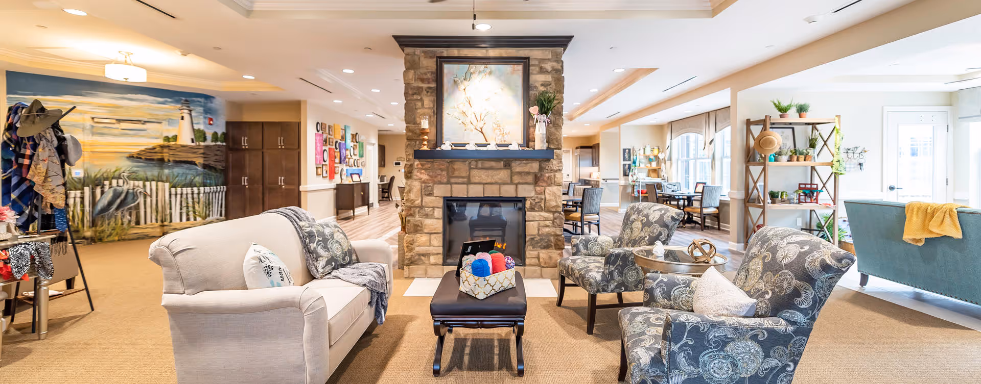 A bright and spacious living room area in a senior living facility featuring a stone fireplace in the center with a painting above it. The room has a beige sofa, two patterned armchairs, a small ottoman with colorful yarn, and a blue couch with a yellow throw. There are large windows letting in natural light, a wooden shelving unit with plants and decor, and a mural of a lighthouse and seagulls on the left wall.