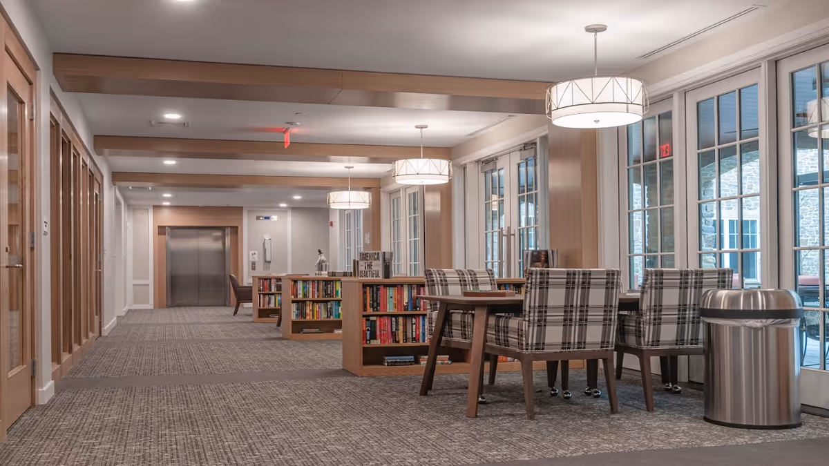 Interior view of a senior living facility hallway with carpeted floors, wooden beams on the ceiling, and large windows along one side. There are bookshelves filled with books and a table with plaid upholstered chairs near the windows. Pendant lights hang from the ceiling, and an elevator is visible at the end of the hallway.