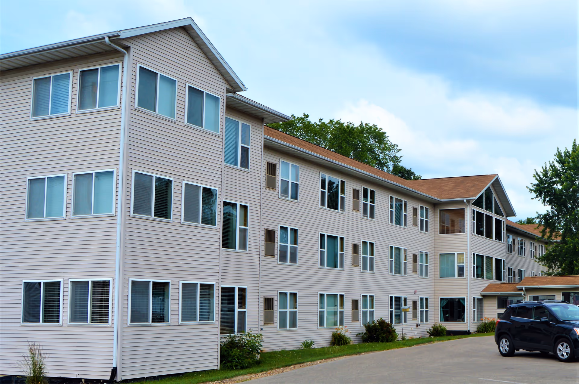 Exterior view of a three-story beige senior living facility building with multiple windows and a brown roof. A black SUV is parked on the paved driveway in front of the building. Trees and a partly cloudy sky are visible in the background.