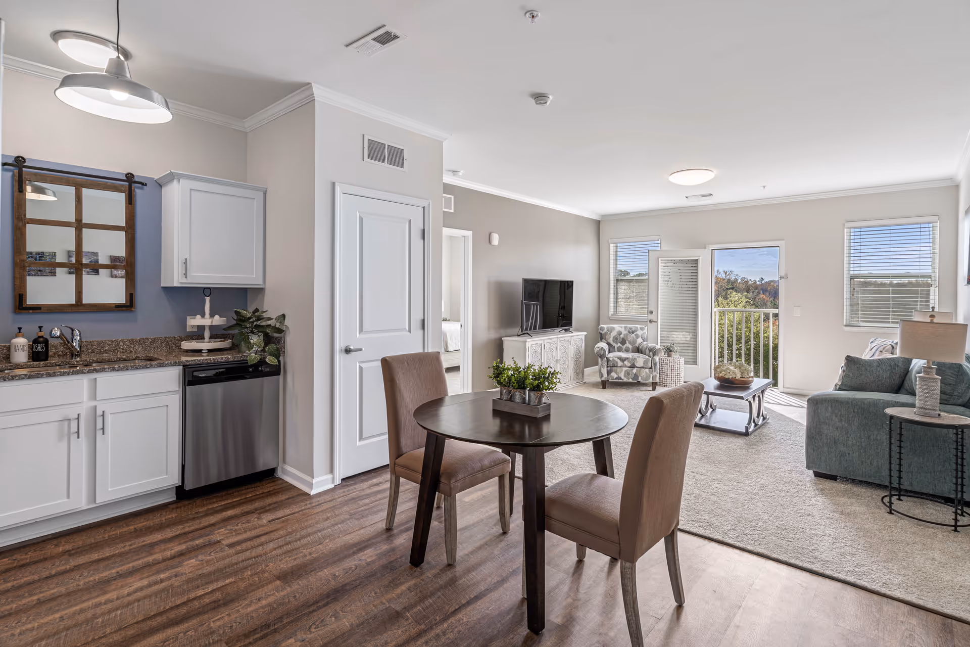 Open-plan apartment interior with a small kitchen, round dining table and chairs, and a furnished living room with couch, armchair, TV, and balcony door.