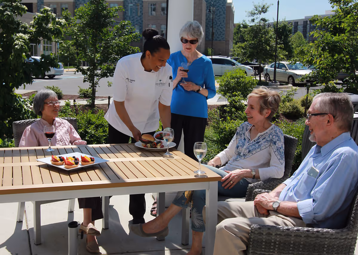 A chef serves plates to four older adults seated around an outdoor patio table with wine glasses and fruit.