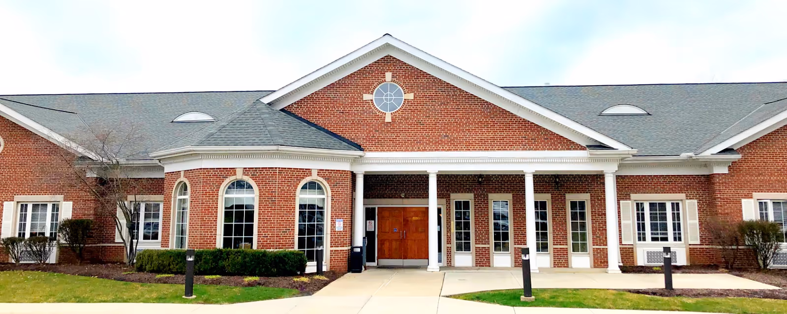Front exterior view of a single-story brick building with a gabled roof, white columns supporting a covered entrance, multiple windows, and a paved walkway leading to double wooden doors.