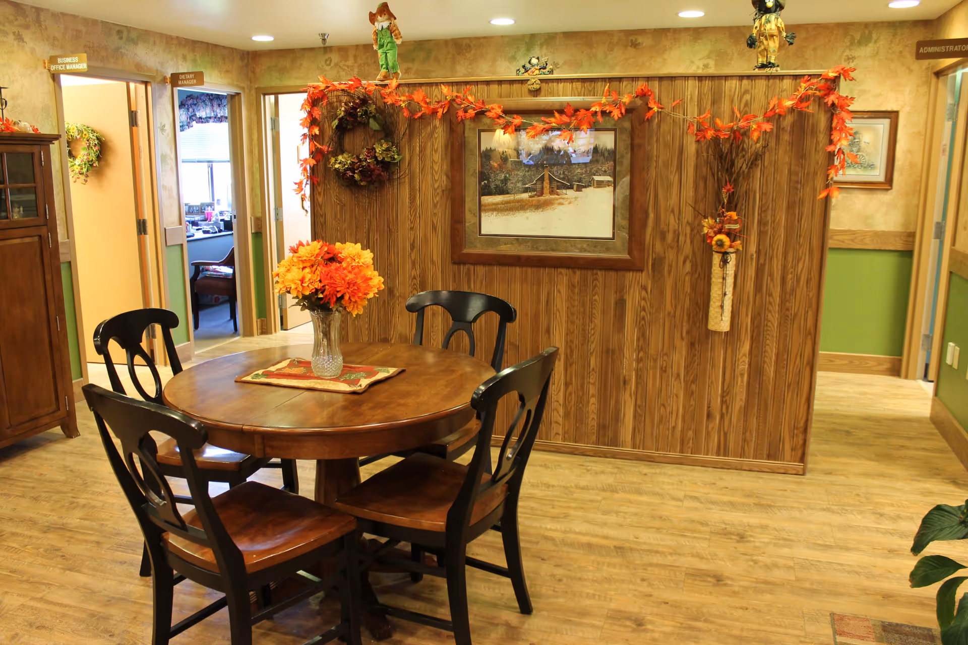 A cozy communal dining area with a round wooden table and four chairs decorated with autumn flowers and wall hangings.