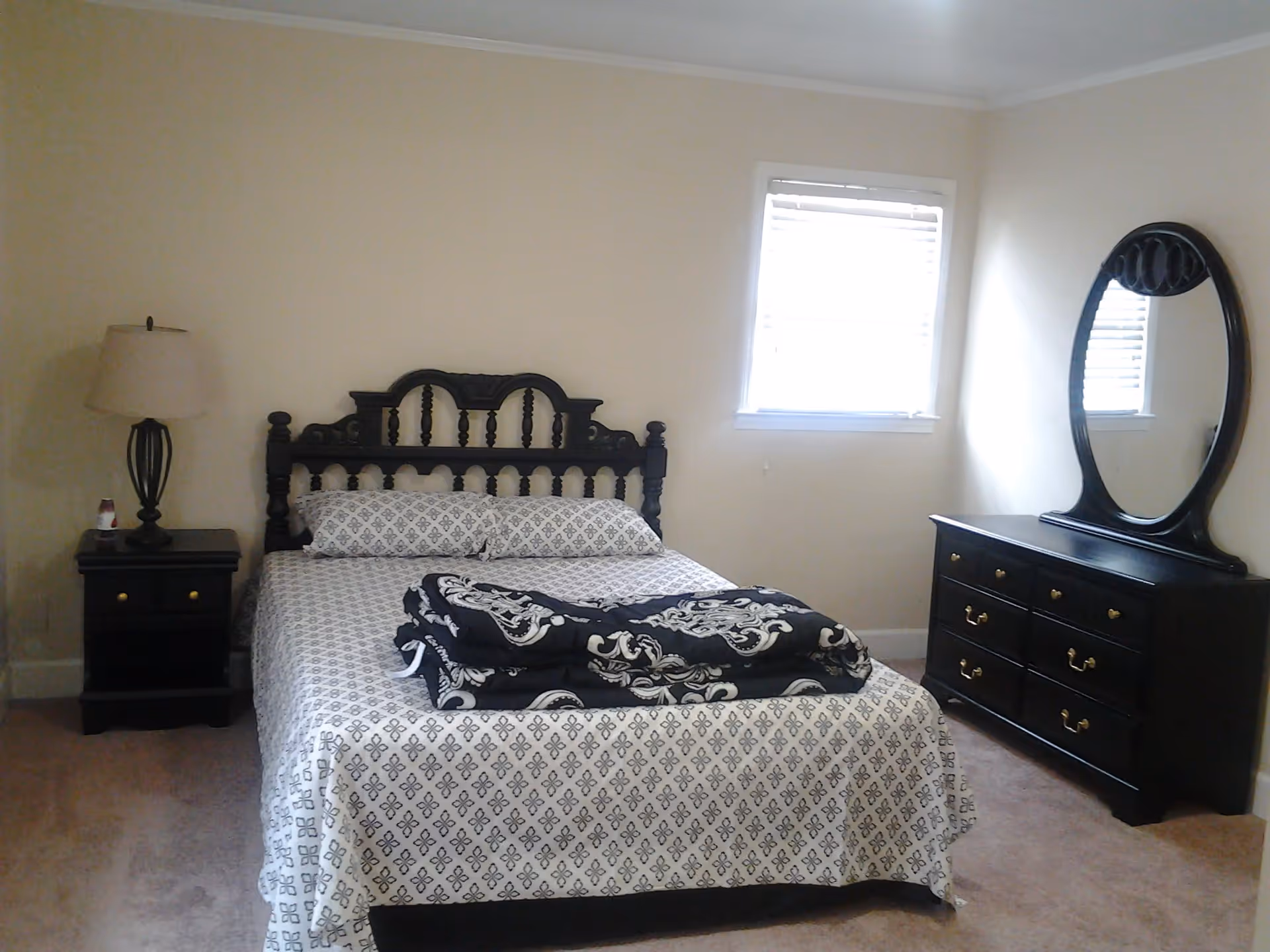 A bedroom with a bed featuring a dark wooden headboard and patterned bedding. There is a folded black and white blanket on the bed. To the left of the bed is a nightstand with a lamp, and to the right is a dresser with a large oval mirror. A window with blinds is on the wall behind the dresser.