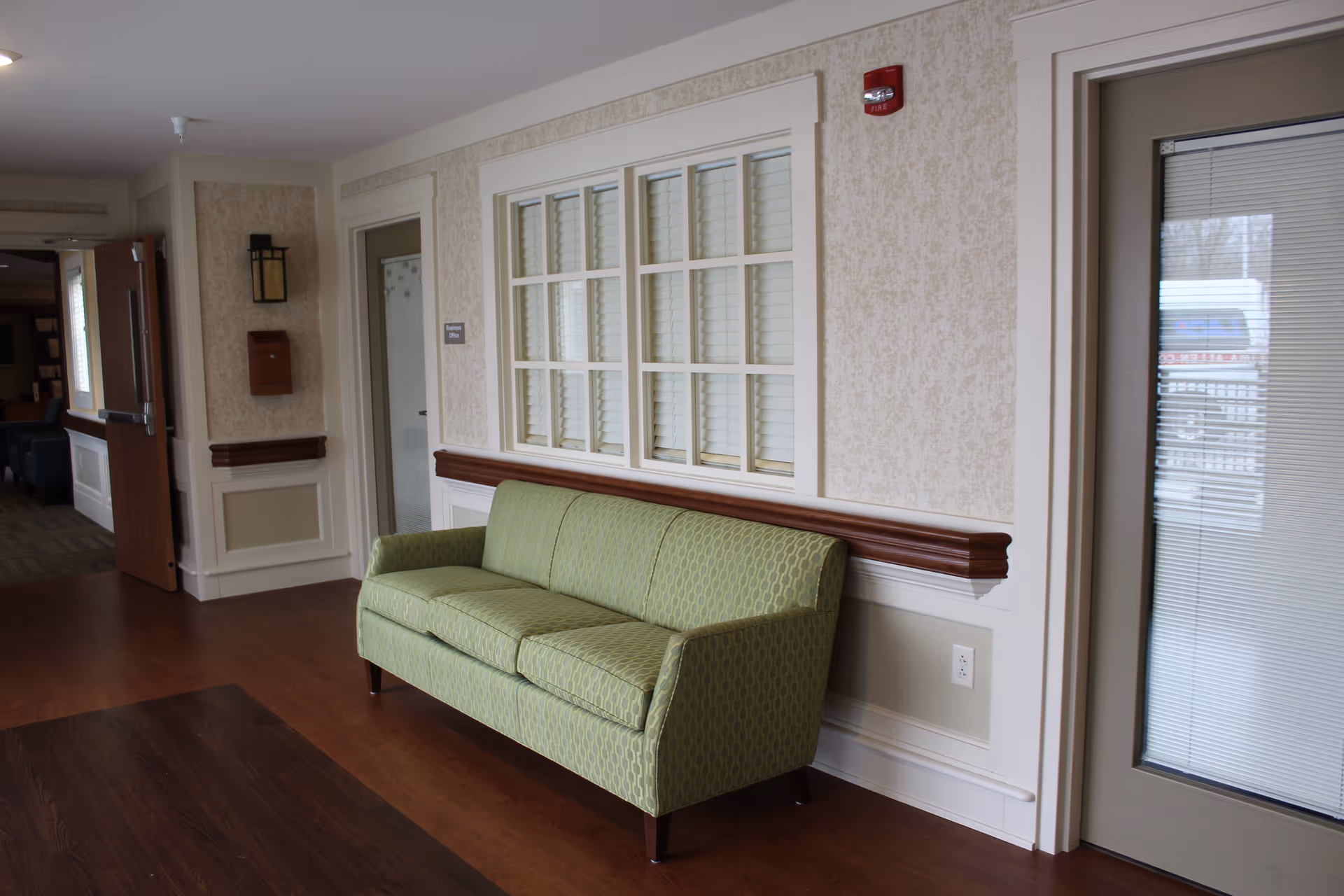 A hallway in a senior living facility with a green patterned sofa against a wall. The wall has a window with white blinds and decorative molding. There is a closed door with a glass panel on the right and an open door leading to another room on the left. The floor is wood, and the walls are light-colored with a textured pattern.