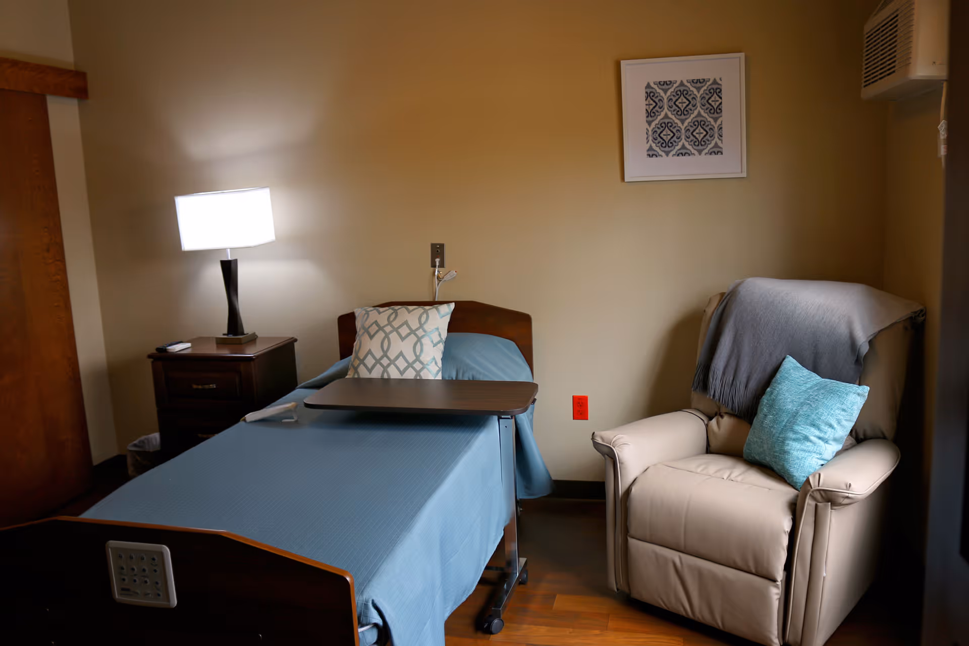 A resident bedroom with a single bed covered in blue bedding, an overbed table, a nightstand with a lamp, and a recliner with pillows.