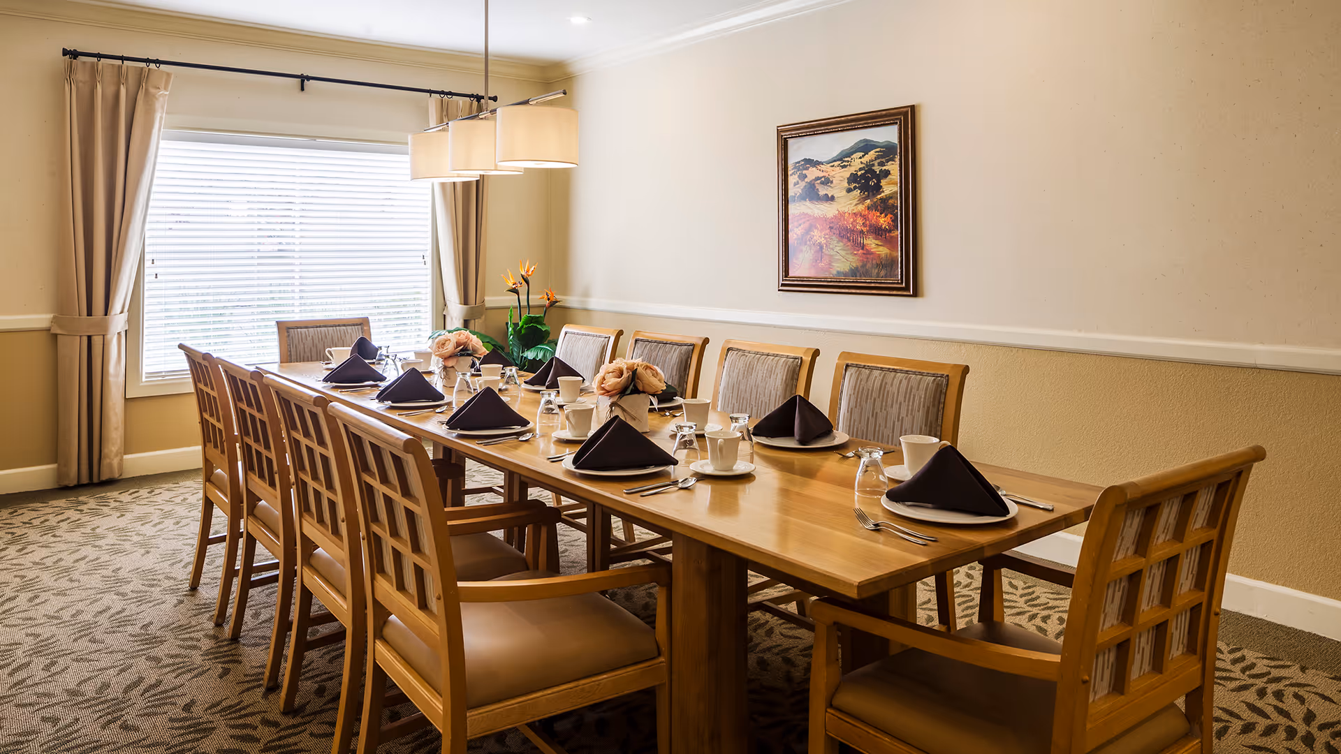 A well-lit dining room with a long wooden table set for twelve people. Each place setting includes a white plate with a folded black napkin, a cup, a glass, and silverware. The room has beige walls, a large window with closed blinds and beige curtains, a hanging light fixture above the table, and a framed landscape painting on the wall.