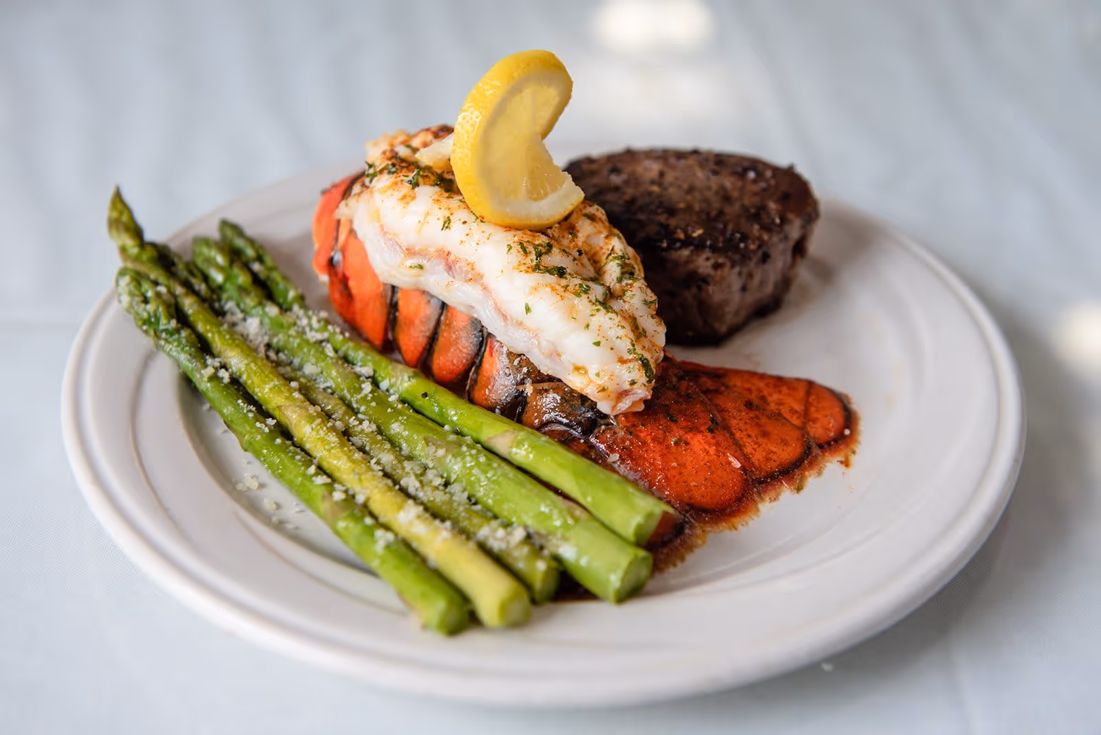 A plated meal consisting of grilled asparagus sprinkled with grated cheese, a lobster tail topped with a lemon wedge, and a piece of grilled steak on a white plate.