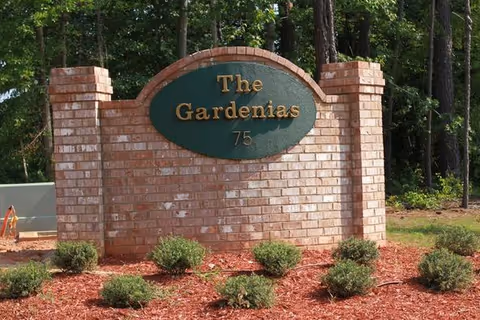 Brick entry sign for The Gardenias with shrubs and trees surrounding it.