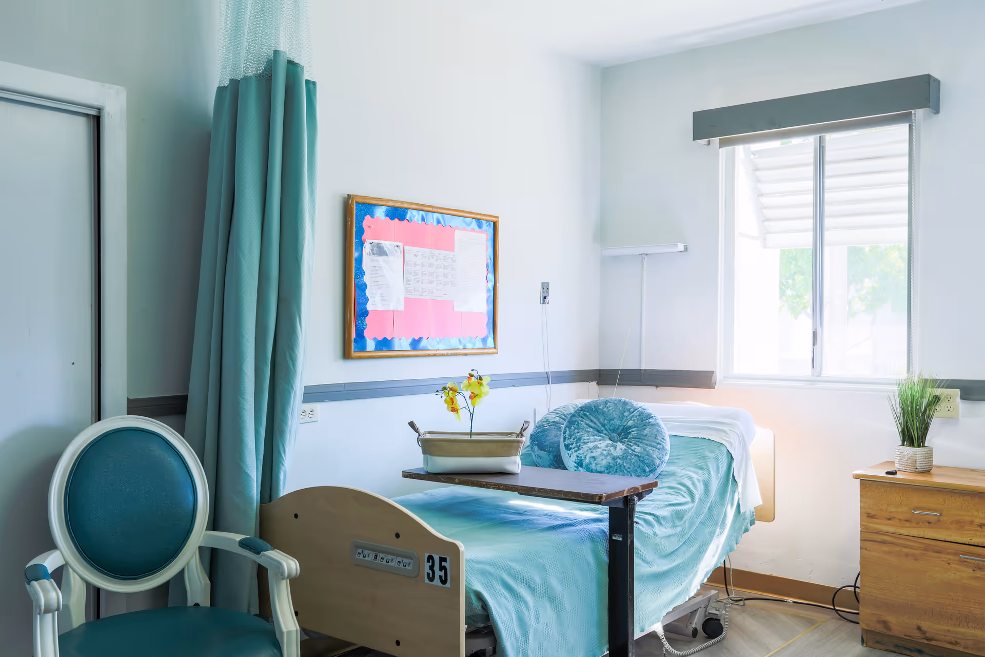 A bright senior care patient bedroom with a single hospital-style bed dressed in teal linens, a matching chair, window, bedside table and bulletin board.