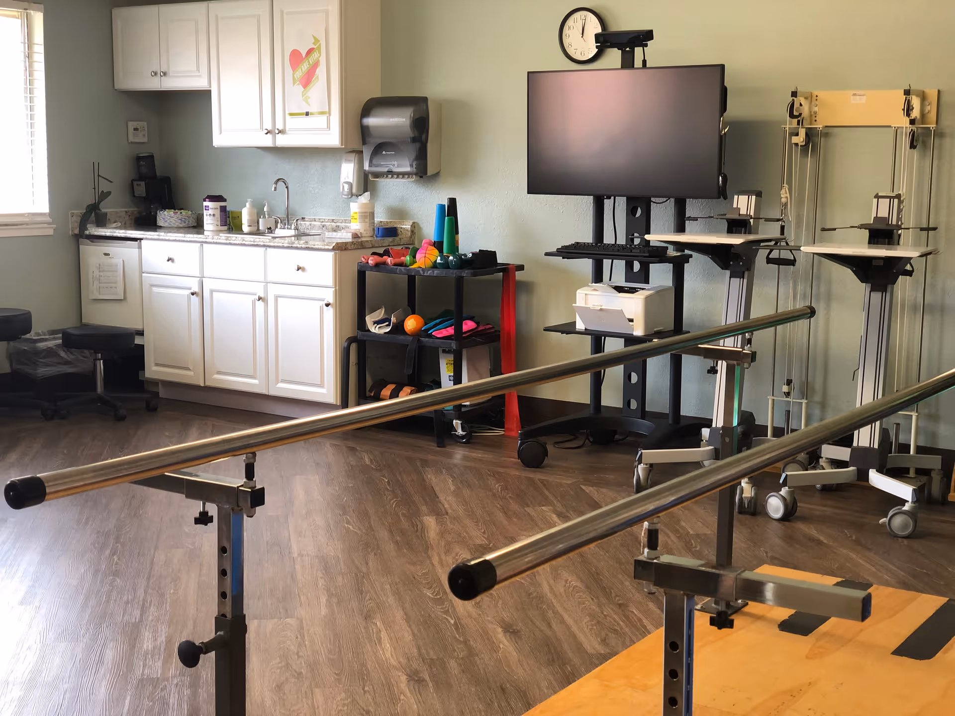 Physical therapy room with parallel walking bars in the foreground, a sink and cabinets on the left, and a monitor and therapy equipment on the right.