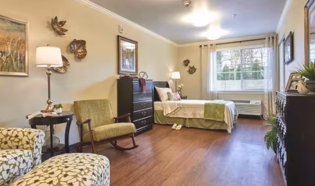 Bright, furnished bedroom with a single bed, dresser, rocking chair, and a large window with blinds.