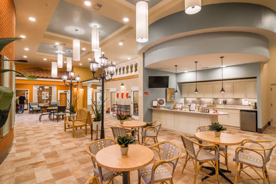 A spacious and well-lit common area in a senior living facility featuring round wooden tables with wicker chairs and small potted plants on each table. To the right, there is a kitchen area with white cabinets, a countertop, pendant lights, and a small refrigerator. The ceiling has recessed lighting and hanging cylindrical light fixtures. In the background, there are additional seating areas with chairs and tables, decorative streetlamp-style lights, and a person walking. The walls have a mix of brick and wood paneling, and there are holiday stockings hanging on a wall near the back.