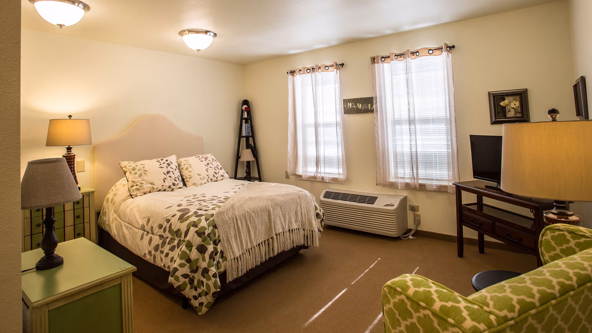 A cozy bedroom with a double bed featuring a leafy patterned comforter and pillows. The room has two windows with sheer curtains, a green armchair with a geometric pattern, two bedside tables with lamps, a small TV on a wooden stand, and a corner shelf with decorative items. The walls are light-colored and the floor is carpeted.