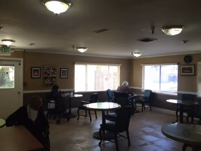 A common area in a senior care facility with several round tables and chairs. Two elderly individuals are seated near the windows, one in a wheelchair and the other in a walker. The room has beige walls, large windows with blinds, and ceiling lights. There are framed pictures on the walls and an exit door on the left side.