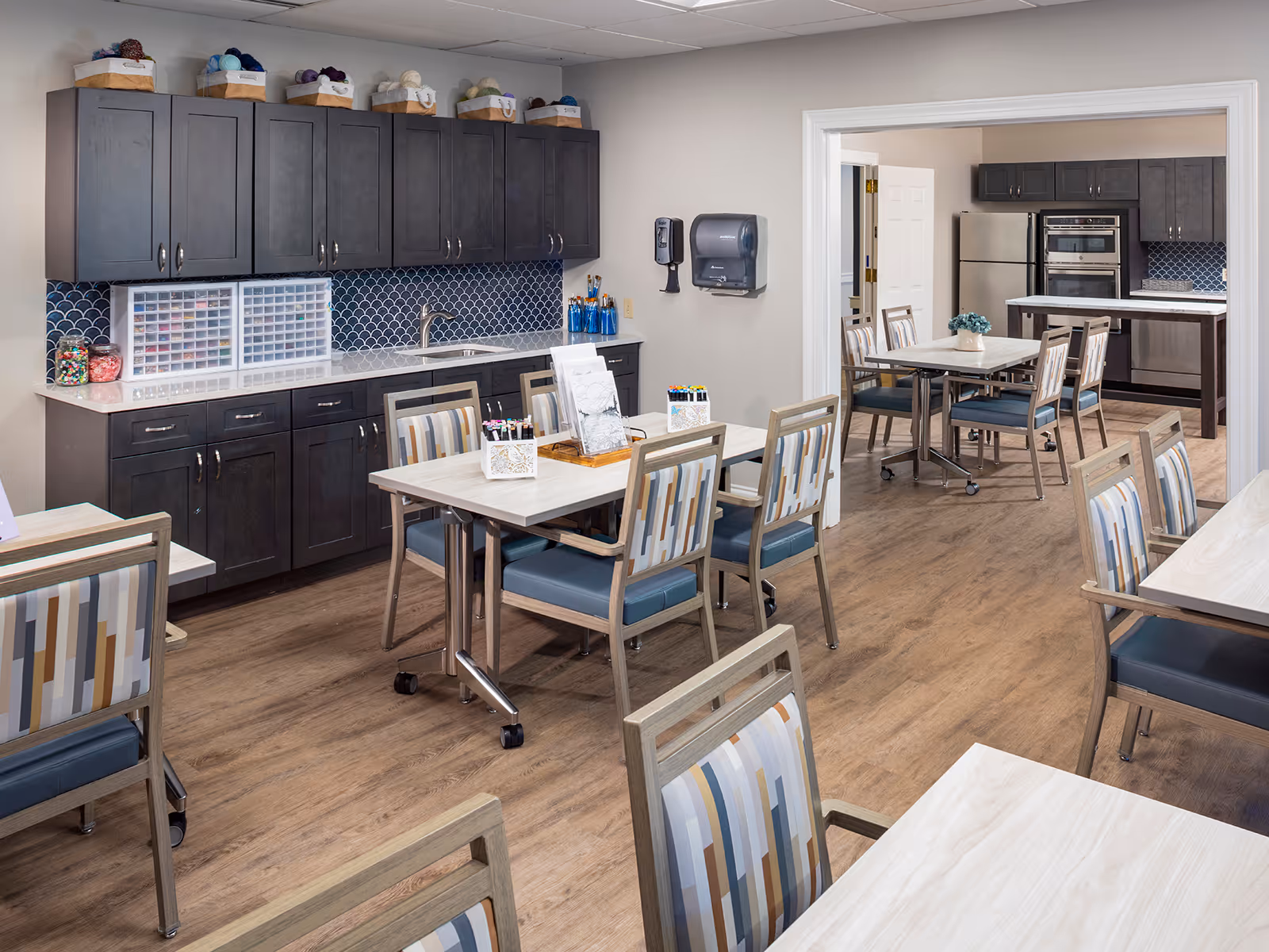 Bright communal dining/activity room with multiple tables and patterned chairs and a kitchenette with dark cabinets and stainless appliances.