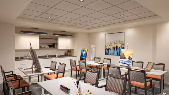 Bright multipurpose activity room with tables and chairs, art easels and supplies, and a kitchenette along the back wall.