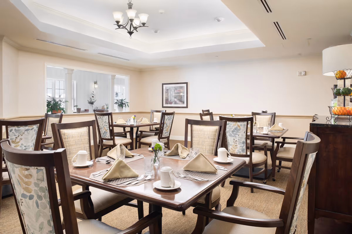 A bright and neatly arranged dining room with several wooden tables and chairs. Each table is set with beige napkins, white cups, glasses, and silverware. The room has light-colored walls, a chandelier, and a framed picture on the wall. There is a window opening to another room with plants visible.