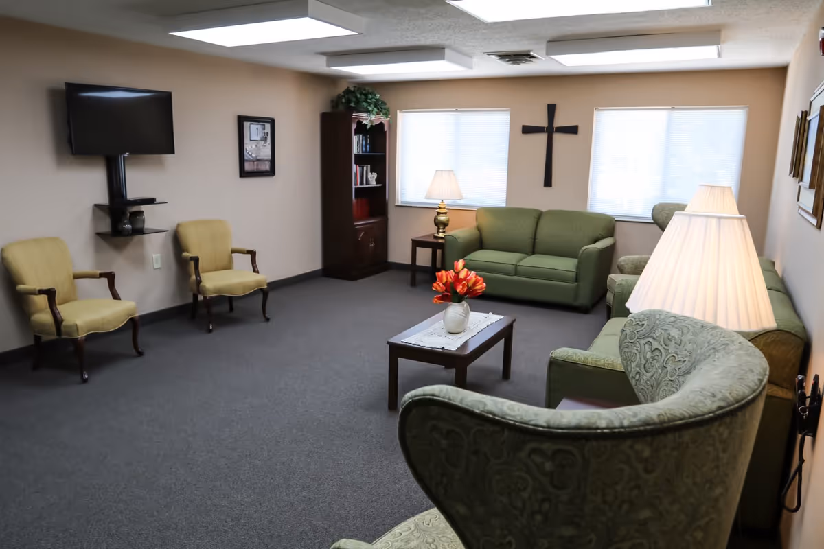 A cozy living room with beige walls and gray carpet. The room features two green sofas, a green patterned armchair, two yellow armchairs, a wooden coffee table with a vase of red flowers, a wooden bookshelf, two table lamps, a wall-mounted TV, and a large cross hanging on the wall between two windows with blinds.