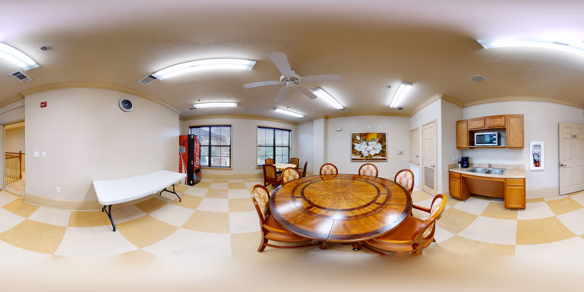A well-lit interior room with a round wooden dining table surrounded by six chairs with patterned upholstery. The room features a small kitchenette area with a microwave, sink, and cabinets. There is a Coca-Cola vending machine near two windows with blinds. The floor has a checkered pattern in beige and light yellow, and the ceiling has fluorescent lights and a white ceiling fan. A large floral painting hangs on the wall above the dining table.