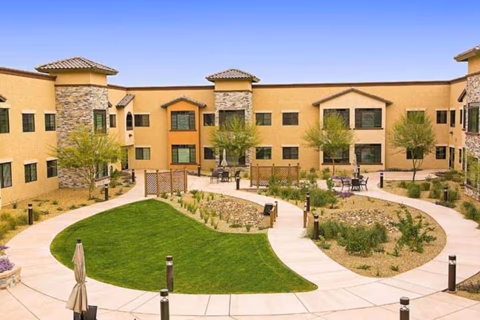 Outdoor courtyard area of a senior living facility with a well-maintained green lawn, paved walkways, small trees, and seating areas with tables and chairs. The building surrounding the courtyard has a beige exterior with stone accents and multiple windows.