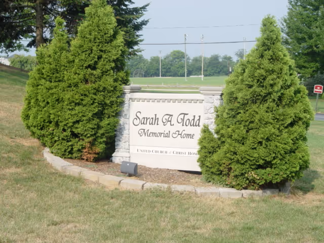 A stone sign for Sarah A Todd Memorial Home surrounded by green grass and two large evergreen trees on either side. The sign is set in a landscaped area with a curved stone border and a small spotlight in front. In the background, there is a grassy field and some trees.
