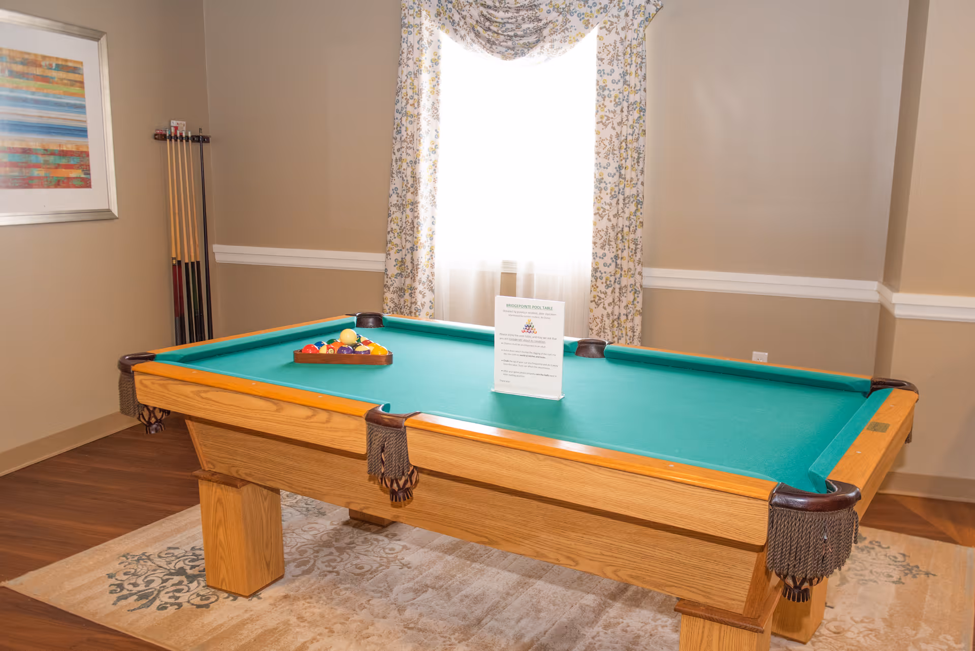 A pool table with green felt in a well-lit game room, cues mounted on the wall and a window with patterned curtains behind it.
