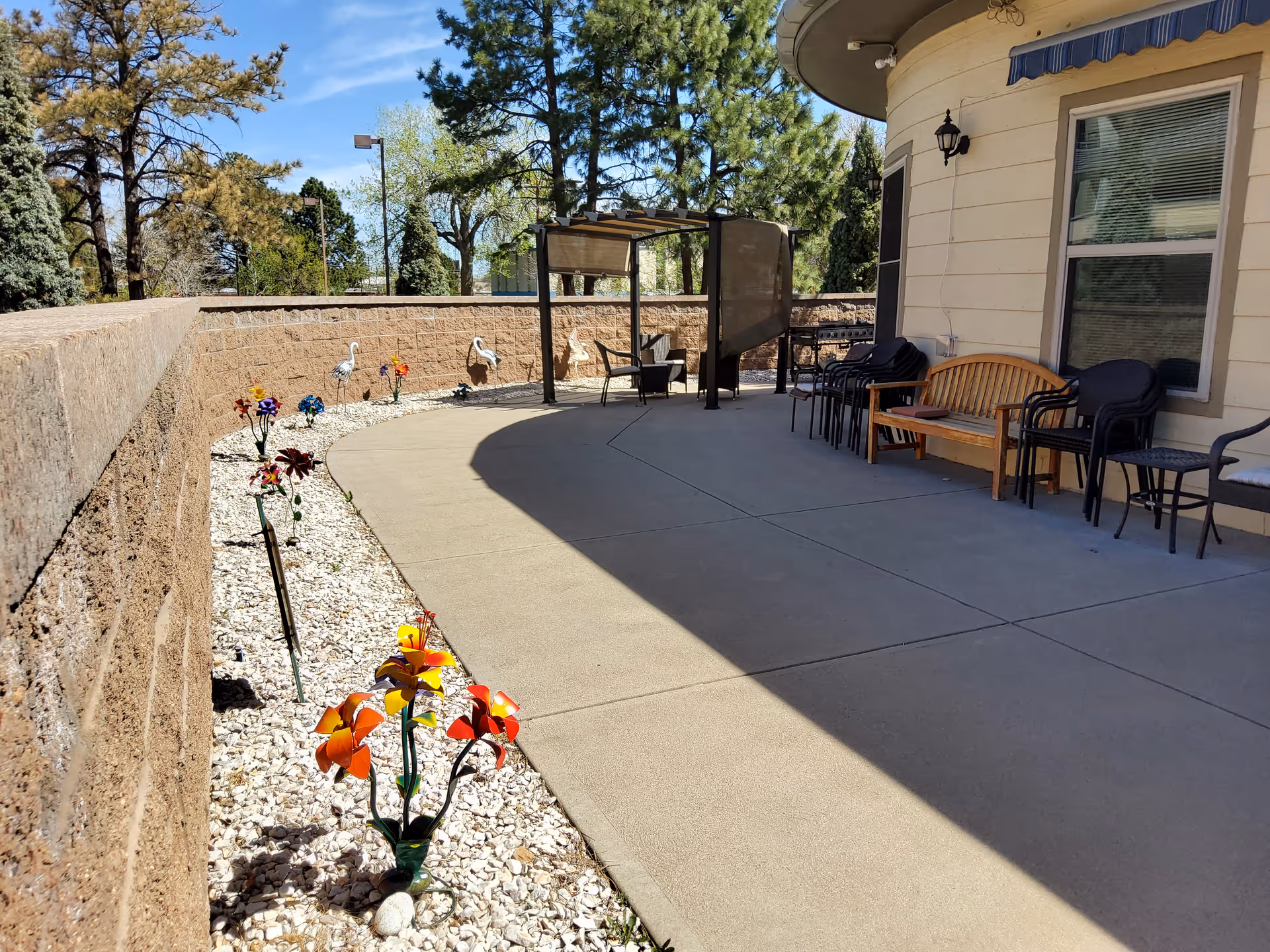 Outdoor patio area at The Granville Assisted Living Center with a curved concrete walkway, a row of colorful metal flower decorations planted in white gravel along a stone retaining wall, several chairs and a wooden bench arranged near the building, and a shaded seating area with a pergola in the background surrounded by trees under a clear blue sky.