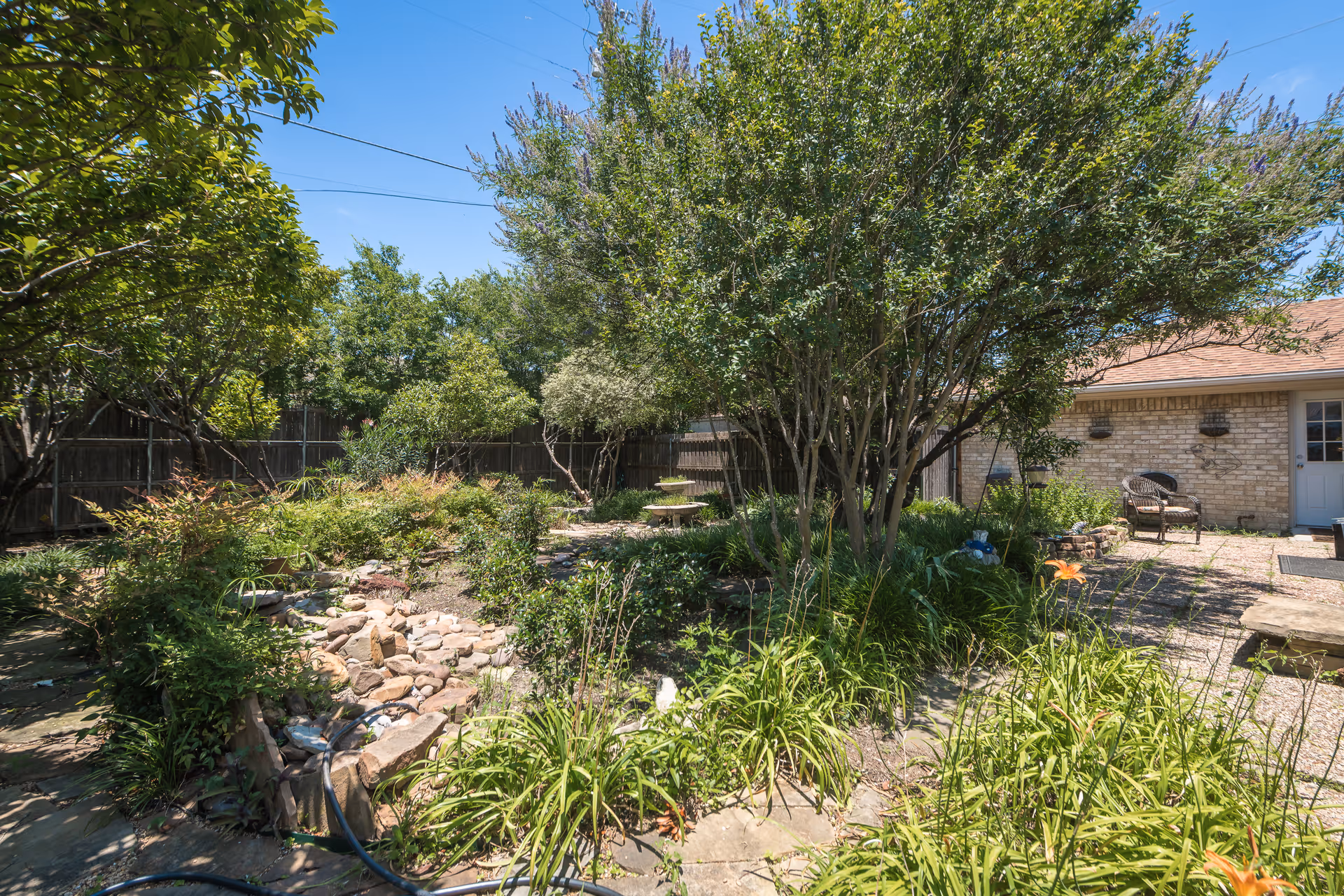 A sunny outdoor garden area with various green plants, bushes, and trees. There is a small stone-lined water feature or dry creek bed in the center. In the background, a brick building with a door and two chairs on a gravel patio is visible. The sky is clear and blue.