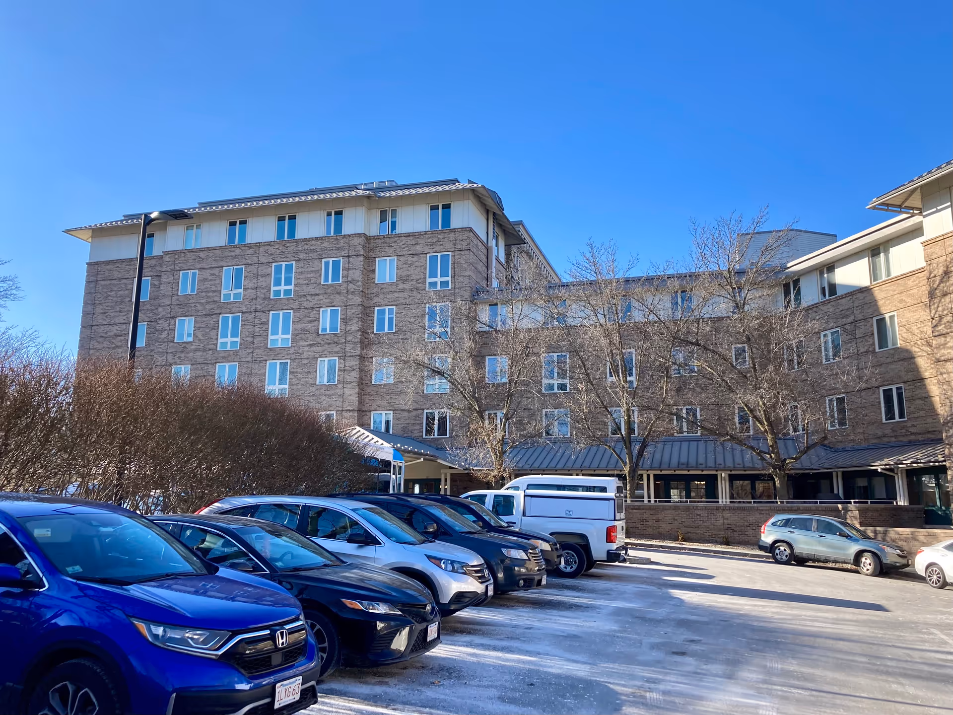 Exterior front view of a multi-story brick senior living building with parked cars in the foreground under a clear blue sky.