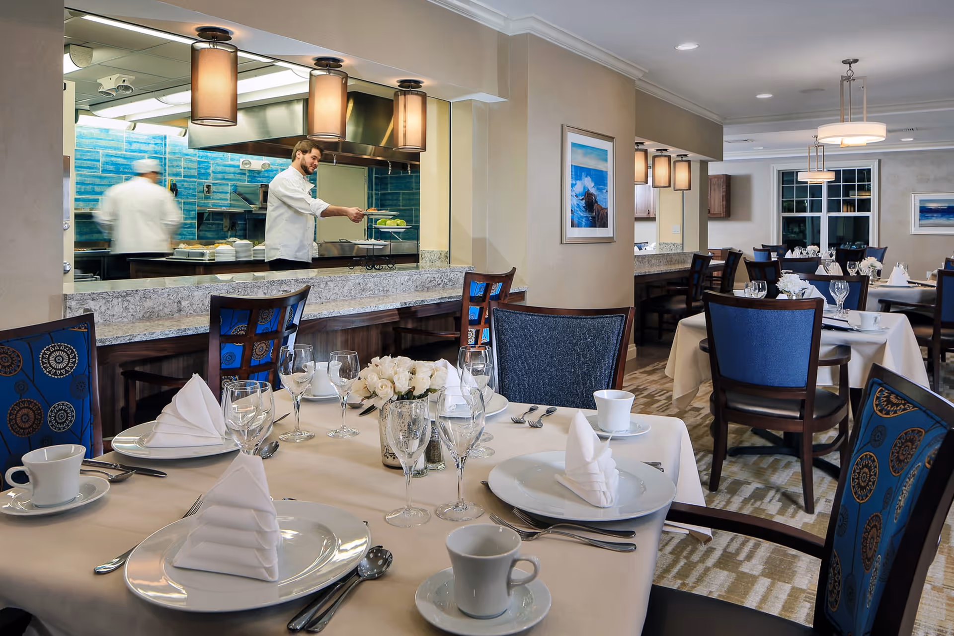 A dining room in a senior living facility with tables set with white tablecloths, folded napkins, glassware, and cups. In the background, chefs are visible through a large serving window with blue tiled walls in the kitchen area. The room has blue and patterned chairs, framed artwork on the walls, and soft lighting fixtures.