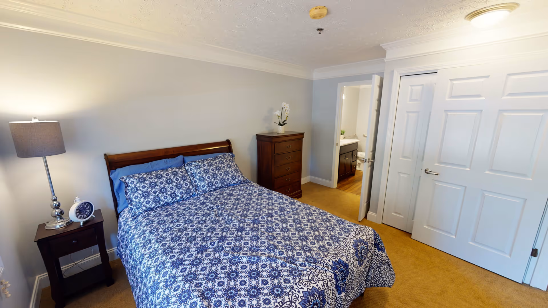 A bedroom with a wooden bed frame and a blue patterned bedspread. There is a nightstand with a lamp and a clock on the left side of the bed. A tall dresser with a small plant on top is against the far wall. The room has beige carpet and light-colored walls. An open door reveals a bathroom with a sink and mirror.