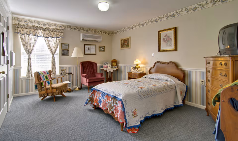 A cozy bedroom with a single bed covered in a patchwork quilt, a wooden headboard, and a wooden dresser with an old-style TV on top. The room has a carpeted floor, floral curtains on the window, a red armchair, a wooden rocking chair with a colorful blanket, and framed pictures on the walls. A small table with a lamp and photos is beside the bed.
