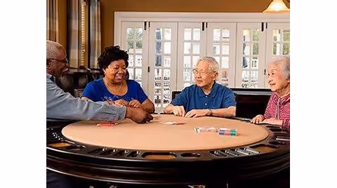 Four elderly individuals sitting around a round table playing a card game in a well-lit room with large windows and French doors in the background.