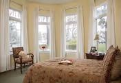 Sunlit bedroom with a bed, armchair, side table, and several tall windows with curtains overlooking greenery.