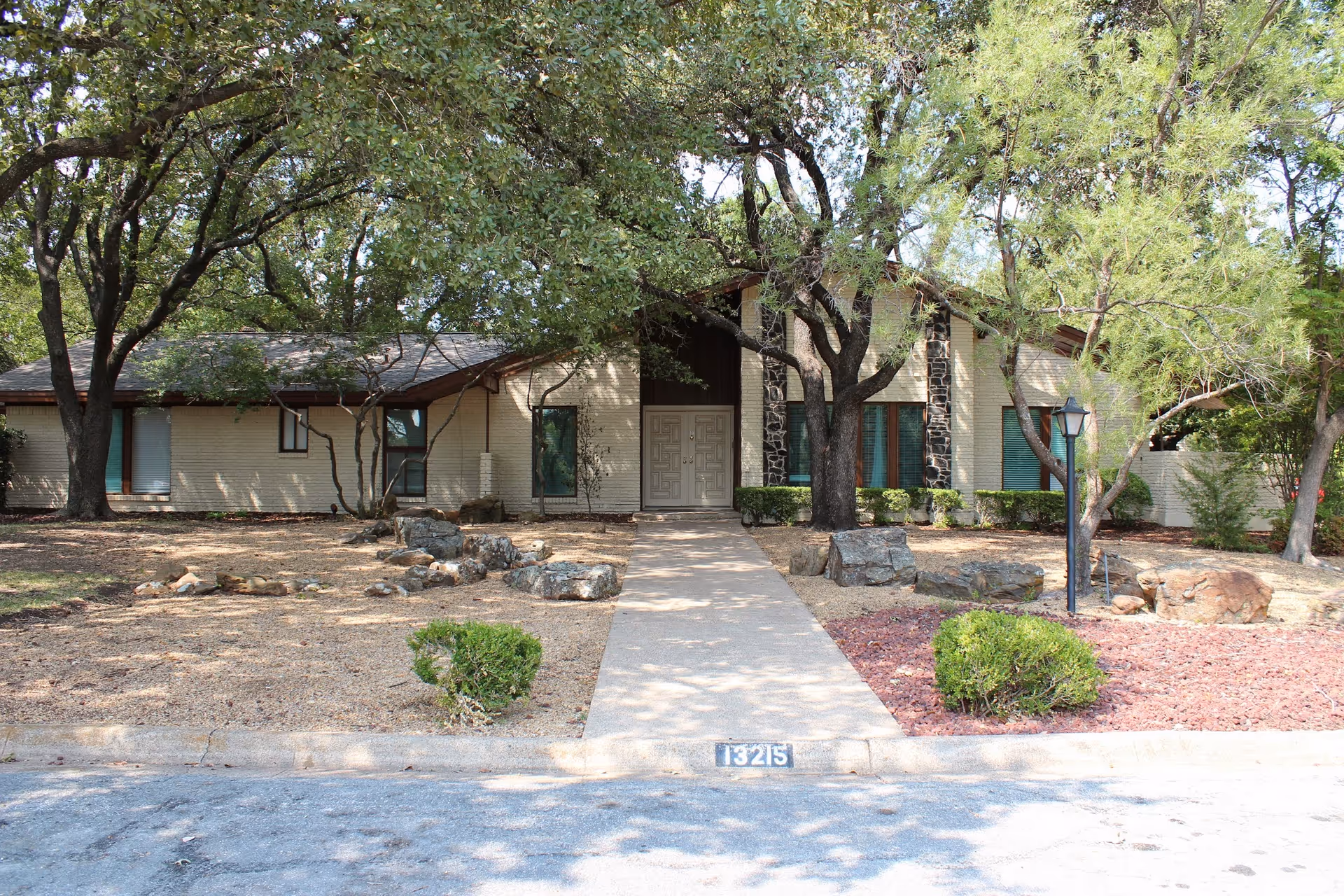 Front exterior view of a single-story building with beige brick walls, a central entrance with double doors, surrounded by trees and landscaping with rocks and small bushes. A concrete pathway leads from the street to the entrance, and the address number 13215 is visible on the curb.