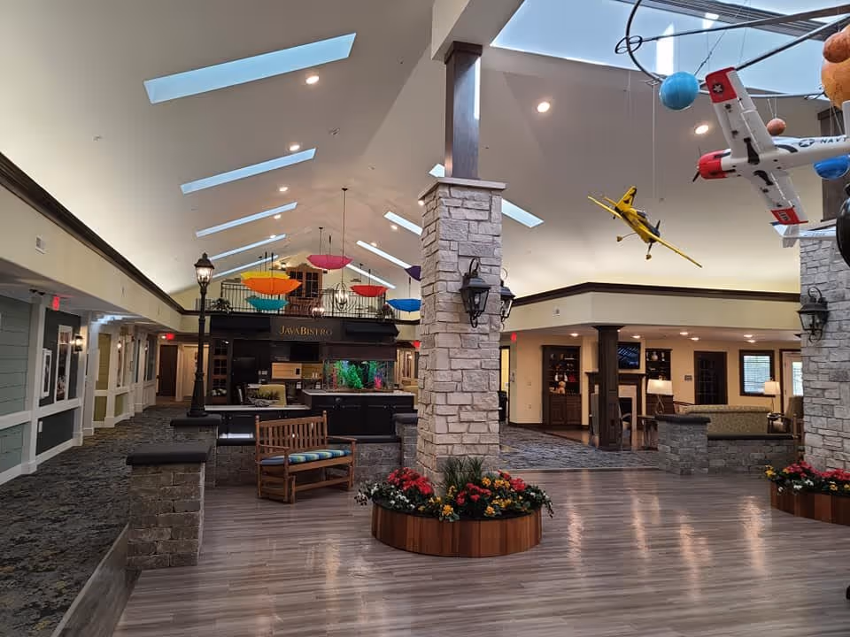Spacious assisted-living interior with a high skylit ceiling, stone pillars, seating areas, hanging colorful umbrellas and model airplanes, and a Java Bistro counter.