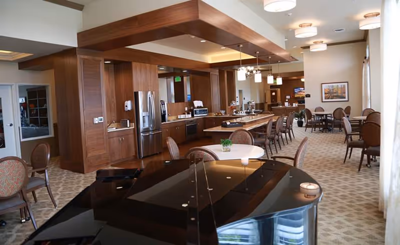 Interior view of a spacious dining and kitchen area in a senior living facility. The room features a long kitchen counter with stainless steel appliances including a refrigerator and microwave. Several round tables with chairs are arranged throughout the carpeted room. A black grand piano is visible in the foreground. The ceiling has modern light fixtures, and large windows with curtains allow natural light to fill the space.