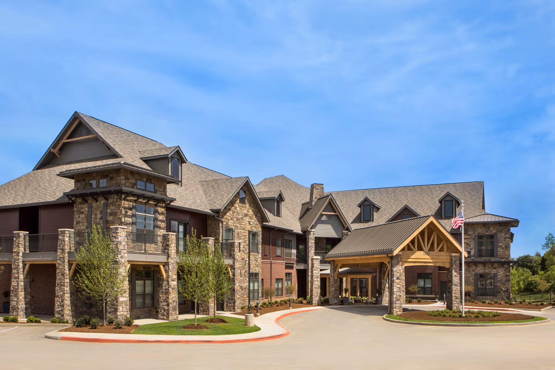 Front exterior of a large stone-and-brick senior living facility with a covered entrance and an American flag.