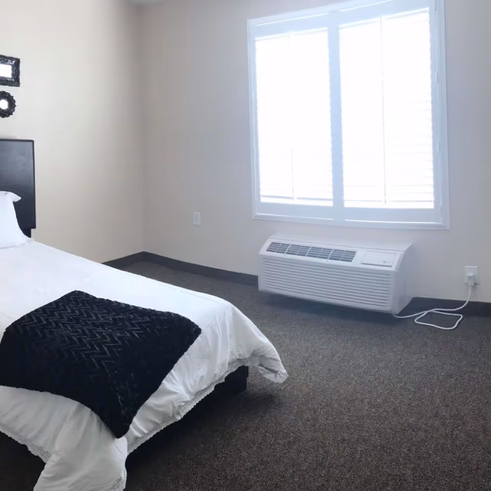 A simple bedroom with a single bed covered in white bedding and a black textured blanket at the foot. The room has beige walls, a window with white blinds, and a white air conditioning unit below the window. The floor is carpeted in a dark color.