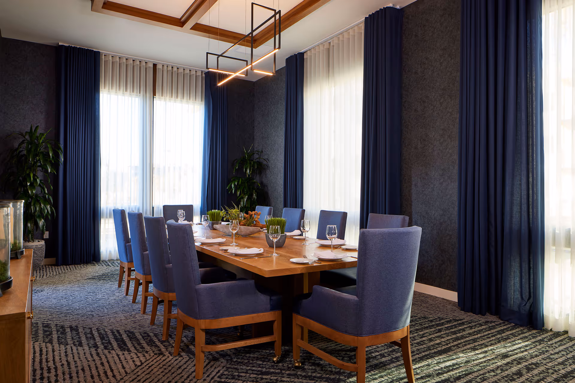 A formal dining room with a large wooden table surrounded by ten blue upholstered chairs. The table is set with white napkins, plates, and wine glasses. The room has large windows with sheer white curtains and dark blue drapes, two green plants in the corners, and a modern rectangular light fixture hanging above the table. The carpet has a patterned design in shades of blue and gray.
