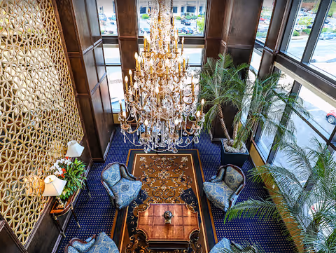 Overhead view of an elegant lobby lounge featuring a large crystal chandelier, four upholstered chairs around a wooden table, patterned carpet, and tall windows with potted palms.