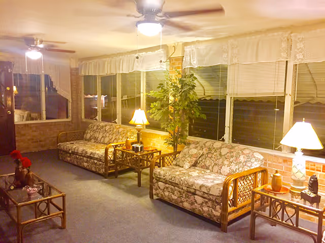A cozy living room area with two floral-patterned sofas, two side tables with lamps, a glass coffee table, a potted plant, and ceiling fans. The room has large windows with blinds and lace curtains, and brick walls.
