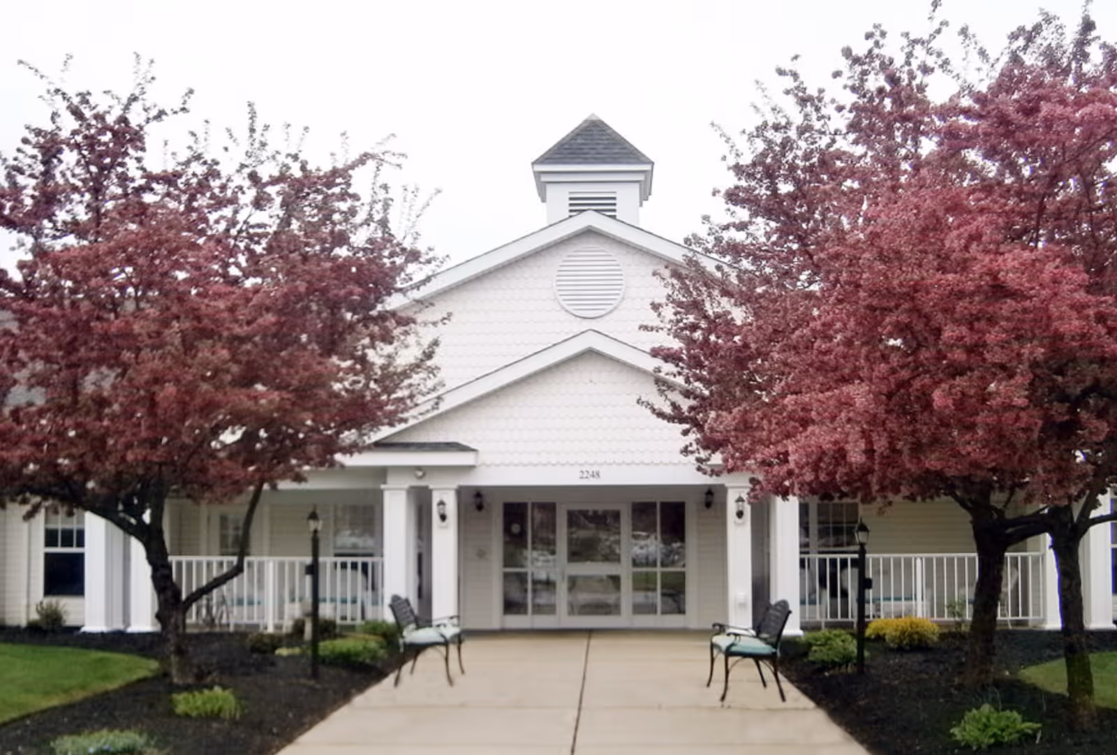 Front entrance of a white senior living building framed by flowering pink trees and benches along the walkway.