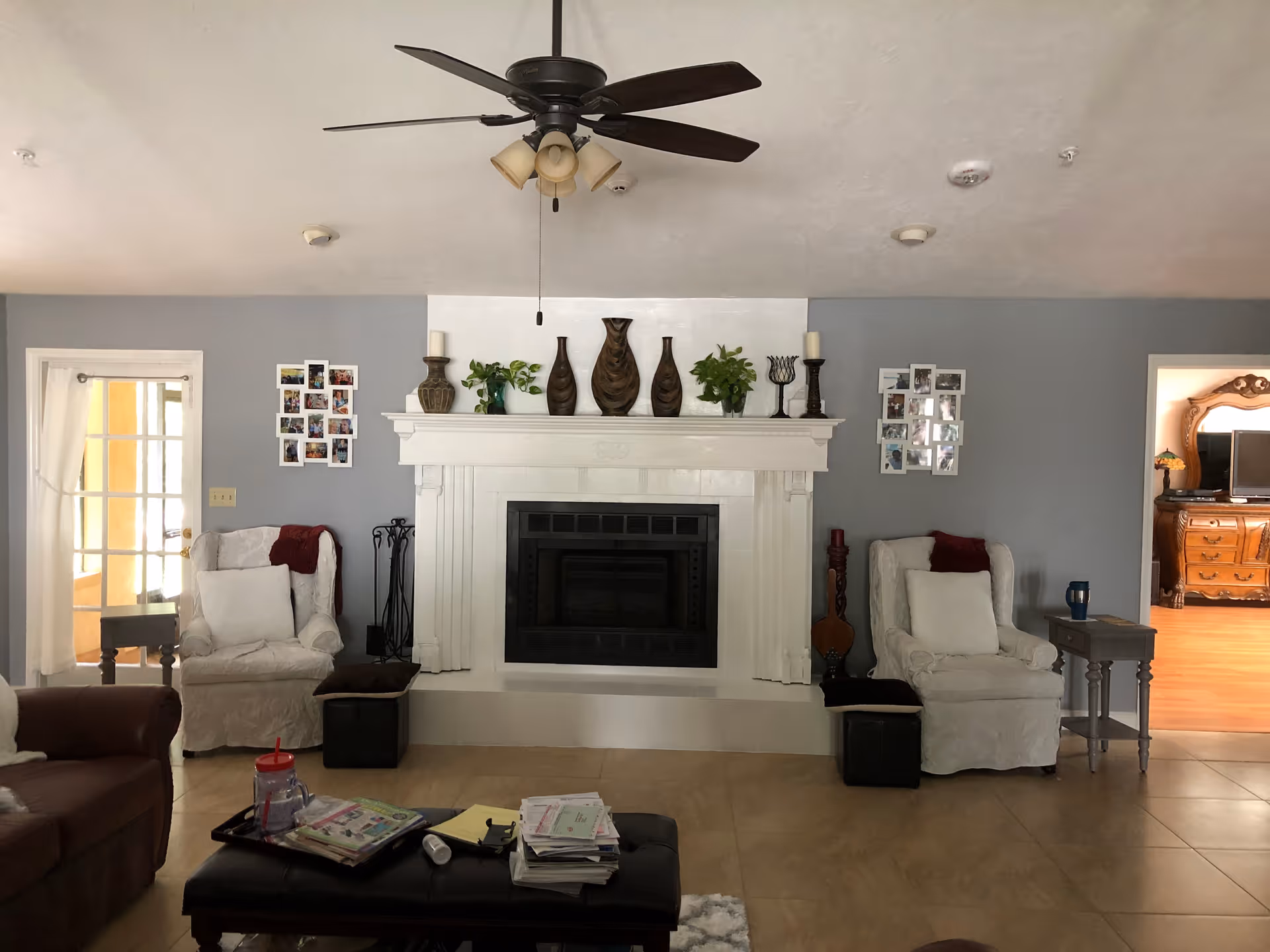 Living room with a central white fireplace flanked by two upholstered armchairs, a ceiling fan, and a coffee table with magazines.