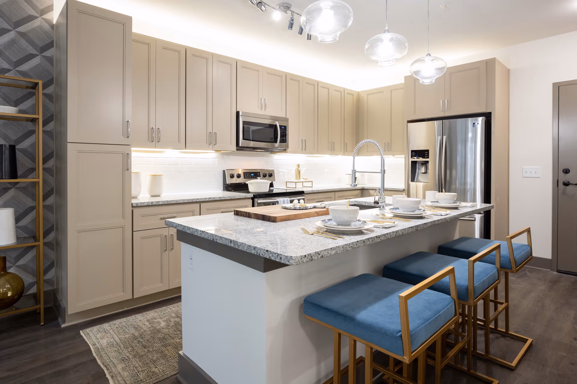 Modern kitchen with beige cabinets, stainless steel appliances including a microwave and refrigerator, a large granite island with a sink and three blue cushioned bar stools, and pendant lights hanging from the ceiling.